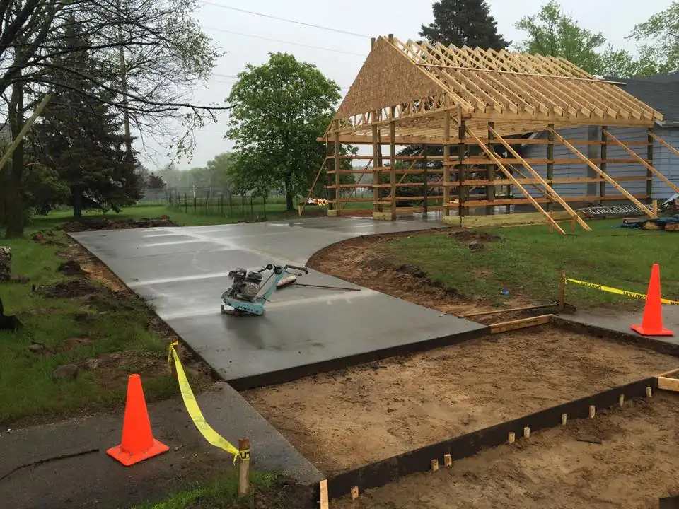 Freshly poured concrete drive with a finishing machine on it, forms and prepared base in front, pole building frame behind