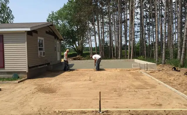Crew levelling a gravel base inside the forms beside a small home, pine trees behind